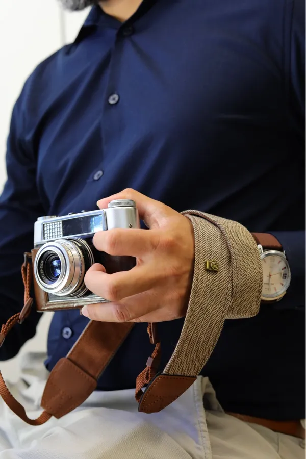 Buyer photo: a man in a navy shirt holding a silver film camera with the camel herringbone strap and its brown leather tabs across his hand