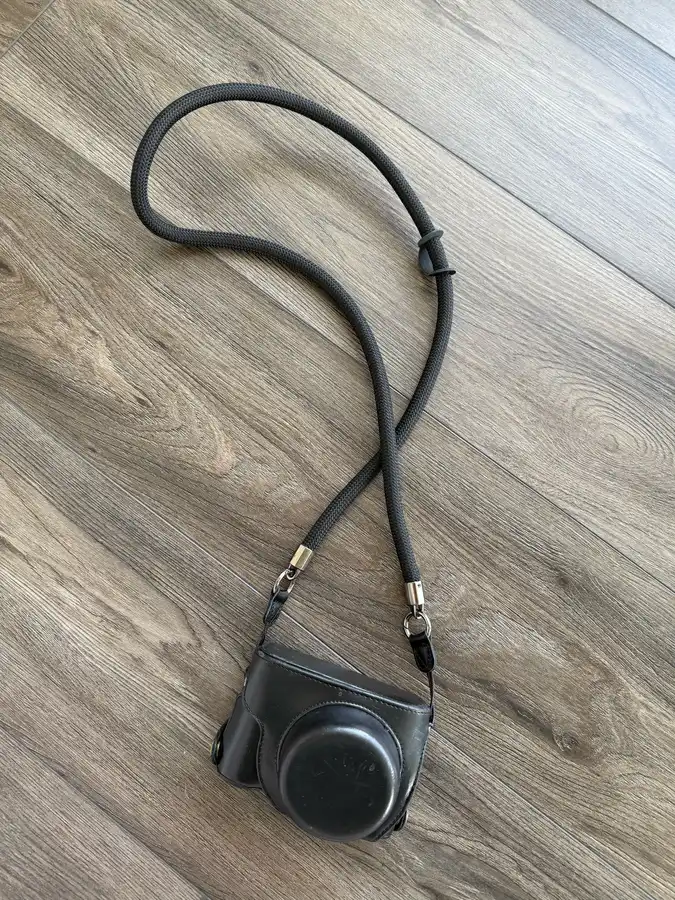 Buyer photo: a dark gray braided rope strap with metal ends laid on a wood floor, clipped to a small black leather camera case