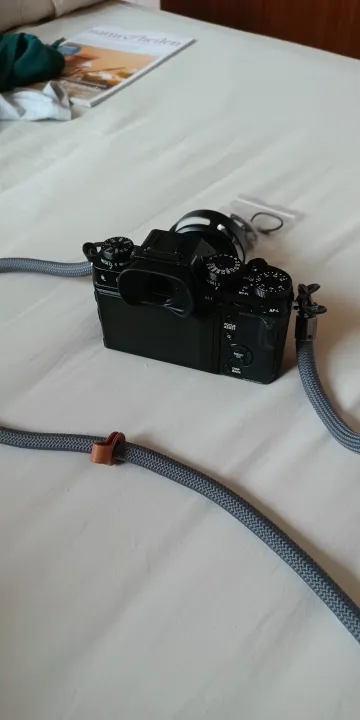 Buyer photo: a black mirrorless camera on a white bed with a gray braided rope strap and a brown leather tab running across the sheet